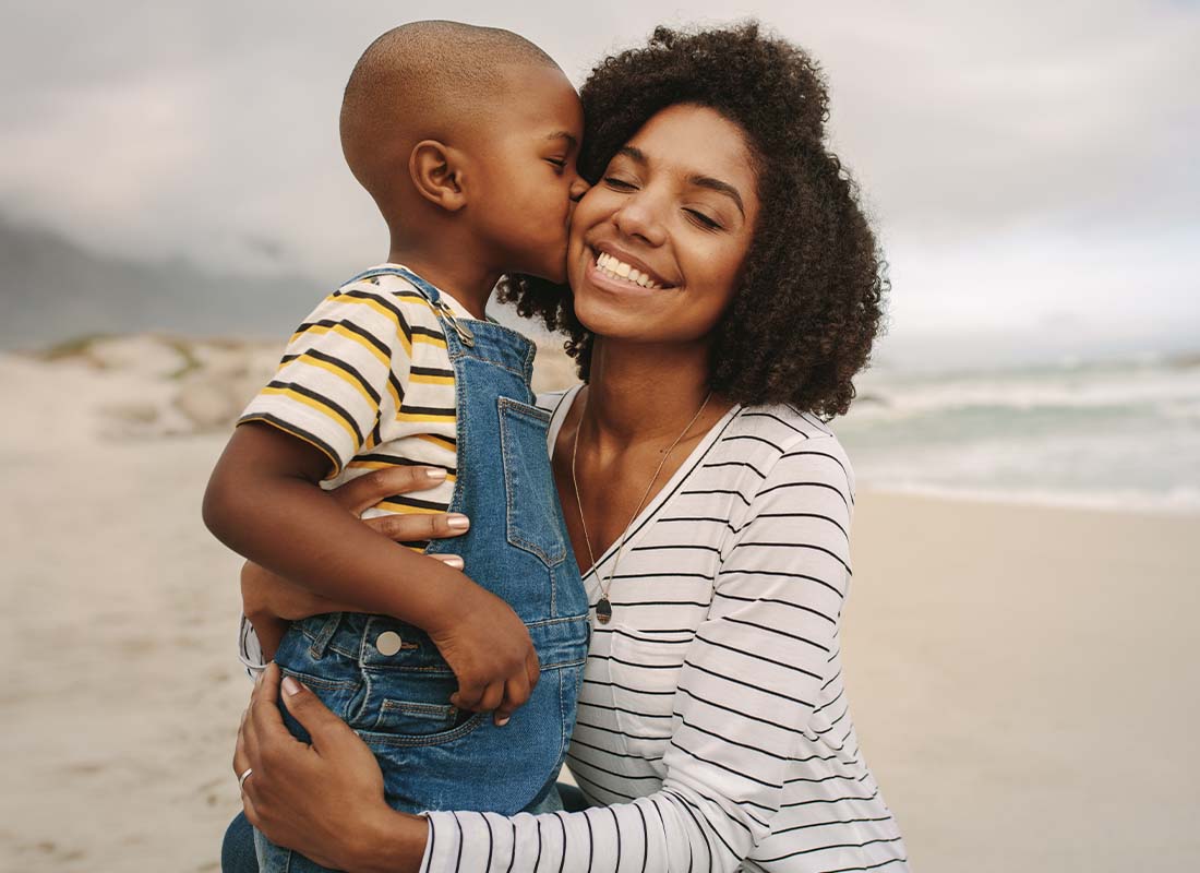 Individual Life Insurance - Young Son Kissing His Smiling Mother While Being Held at the Beach on a Cloudy Day